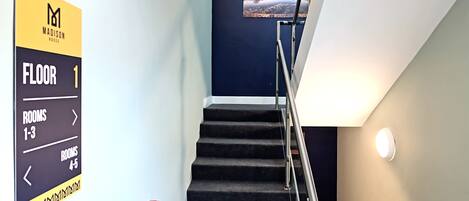 Interior de un hotel con una escalera que conduce al primer piso, señalización direccional para las habitaciones 1-3 y 4-5, y un letrero de circuito cerrado de televisión.