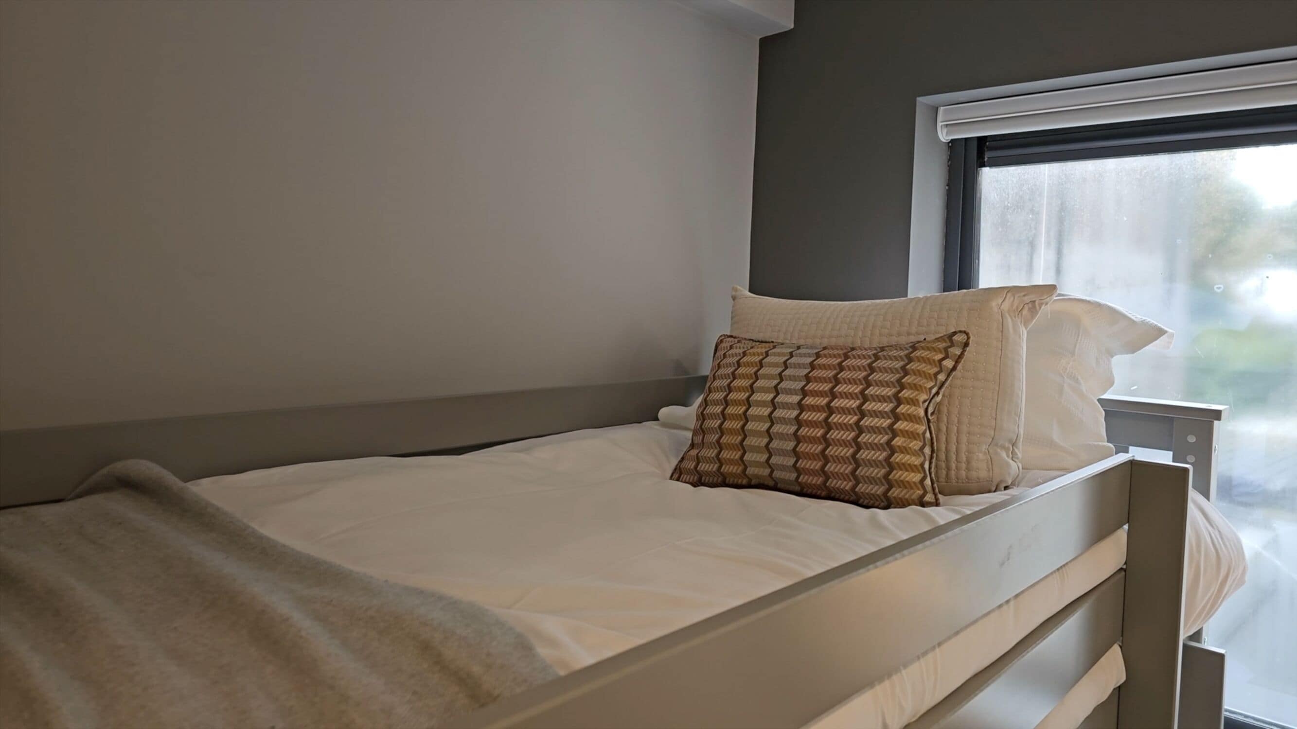 Una cama individual con un cojín estampado y una manta blanca en una habitación con ventana.