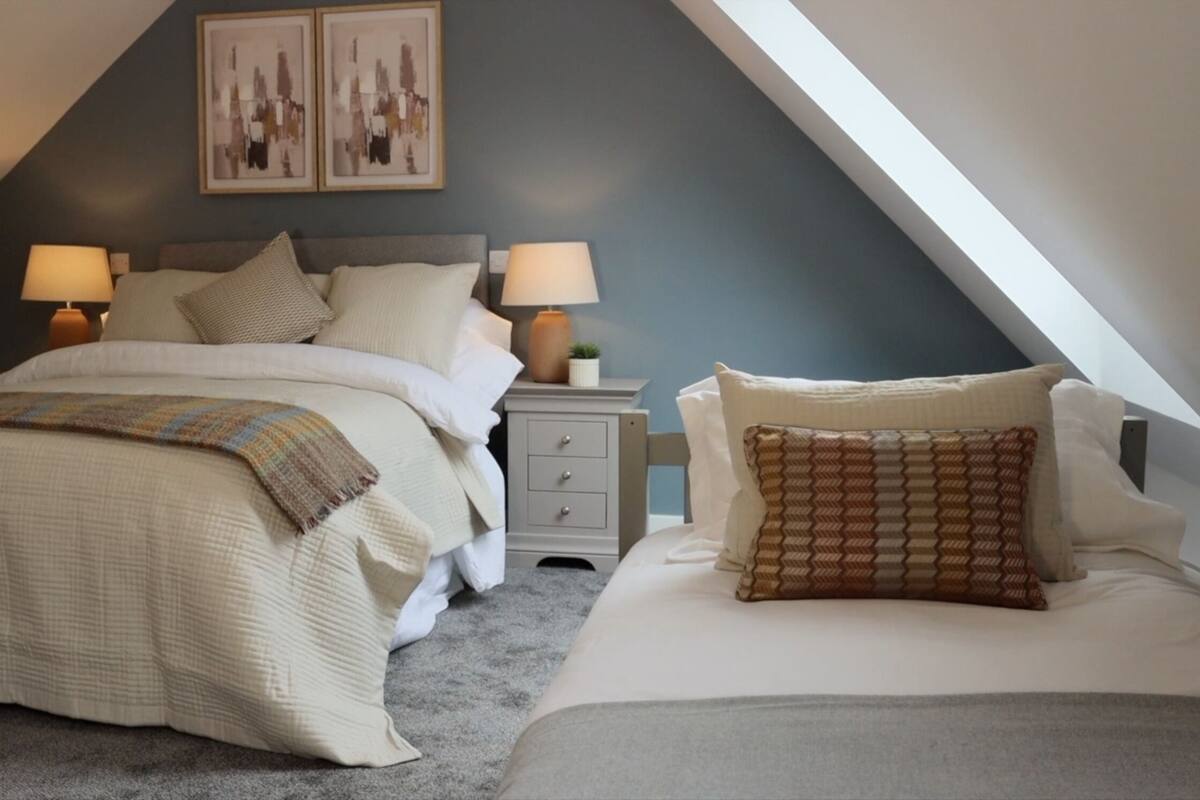 Une chambre avec un plafond en pente, un lit recouvert d’une couverture matelassée, deux tables de chevet avec des lampes et une petite table de nuit ornée d’une plante.