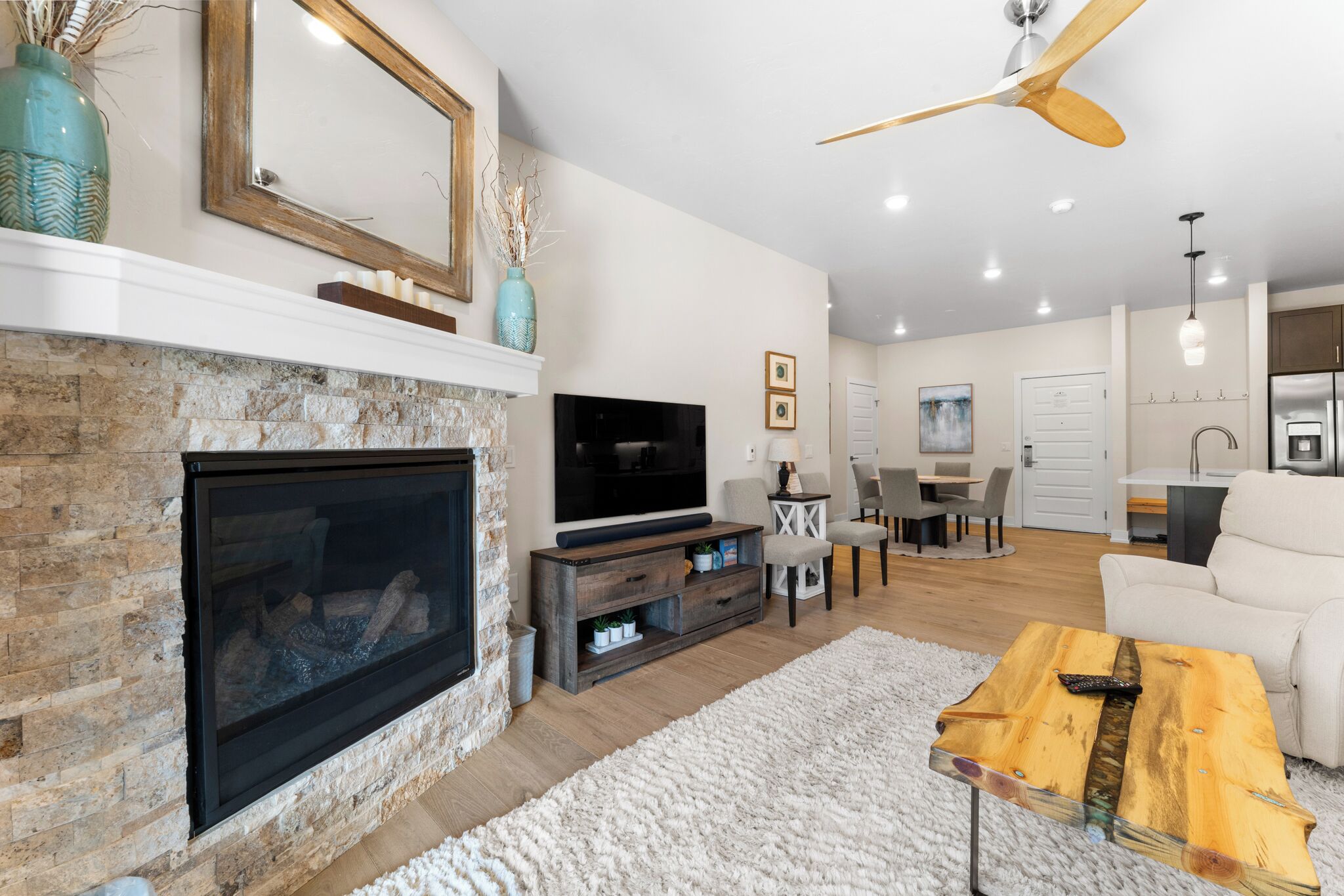 A modern living room with a stone fireplace, a flat-screen TV, a wooden coffee table, and a ceiling fan.