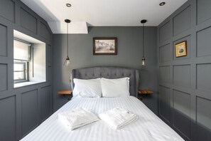Ein Schlafzimmer mit grauem, gepolsterten Kopfteil, zwei weißen Kissen, einem ordentlich bezogenen Bett mit weißen Leinen und einem gerahmten Bild an der Wand.
