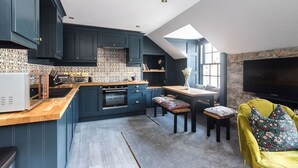 A modern kitchen with dark blue cabinets, a wooden countertop, and a built-in oven. There is a dining area with a bench and a table, and a television mounted on the wall. - Boutique Castle View 3 Bed, 1 Bath Apartment

​ (Edinburgh)
