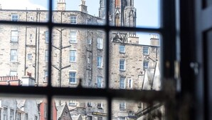 A view through a window with a grid pattern, featuring a street scene with buildings, a car, and a statue in the foreground. - Boutique Castle View 3 Bed, 1 Bath Apartment

​ (Edinburgh)