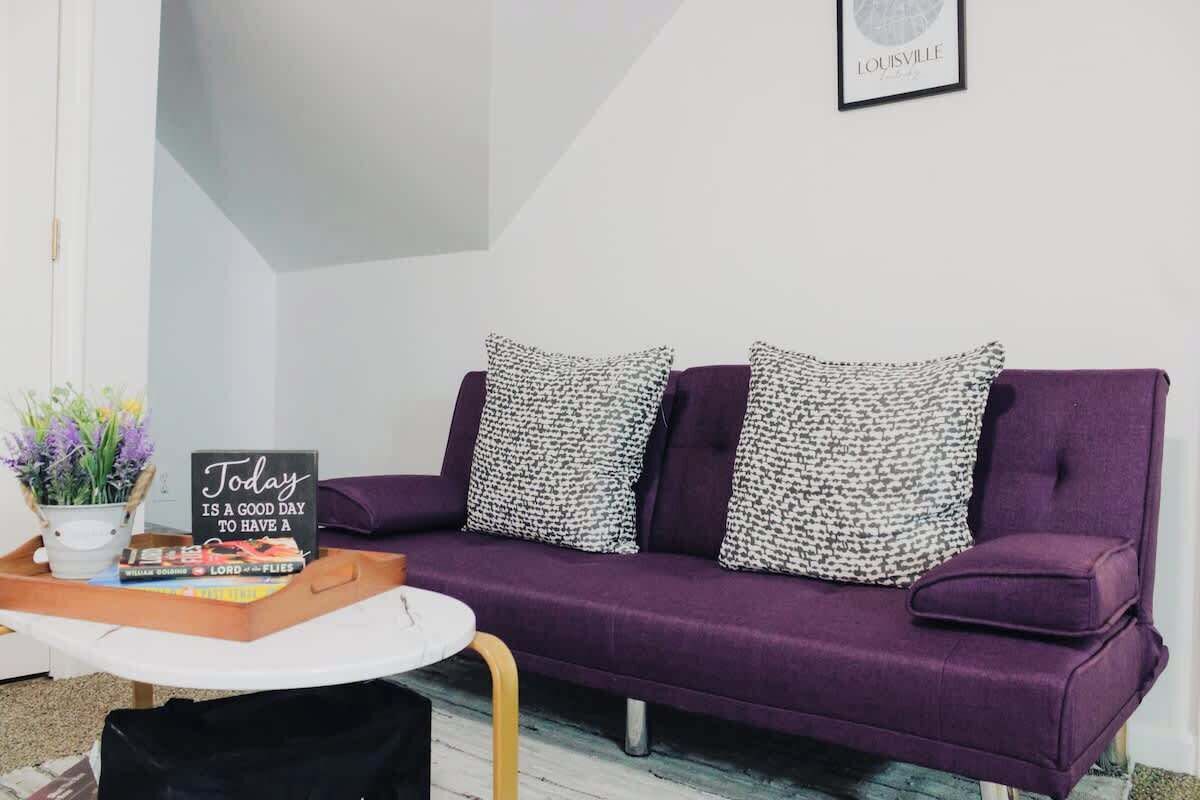 A purple sofa with patterned cushions, a small table with a sign, and a framed picture on the wall.