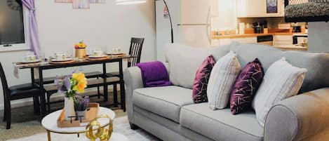 A living room with a grey sofa, a dining table with chairs, a small round table with a vase, and a kitchen area in the background.