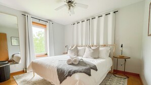 Une chambre à coucher bien aménagée, avec un grand lit, un ventilateur de plafond, une table de chevet avec une lampe et une vue sur l’extérieur par une fenêtre.
