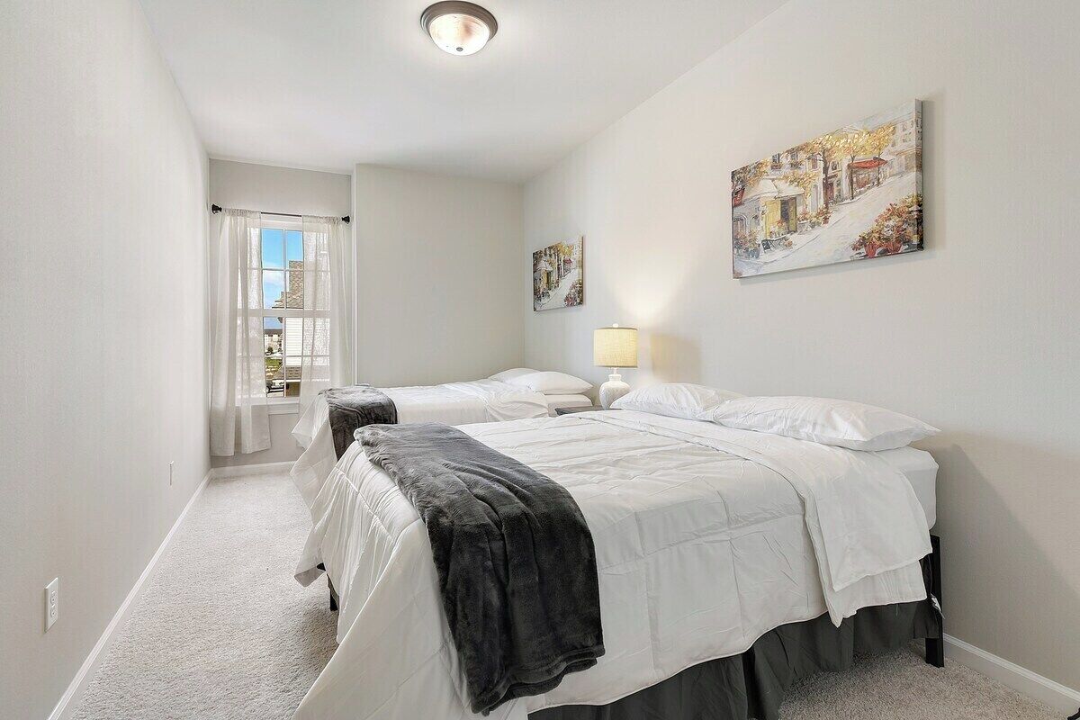 A bedroom with two beds, a window with curtains, a lamp, and artwork on the wall.