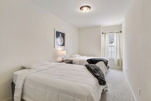 Un dormitorio con dos camas, una mesita de noche, una lámpara, un cuadro en la pared y una ventana con cortinas.