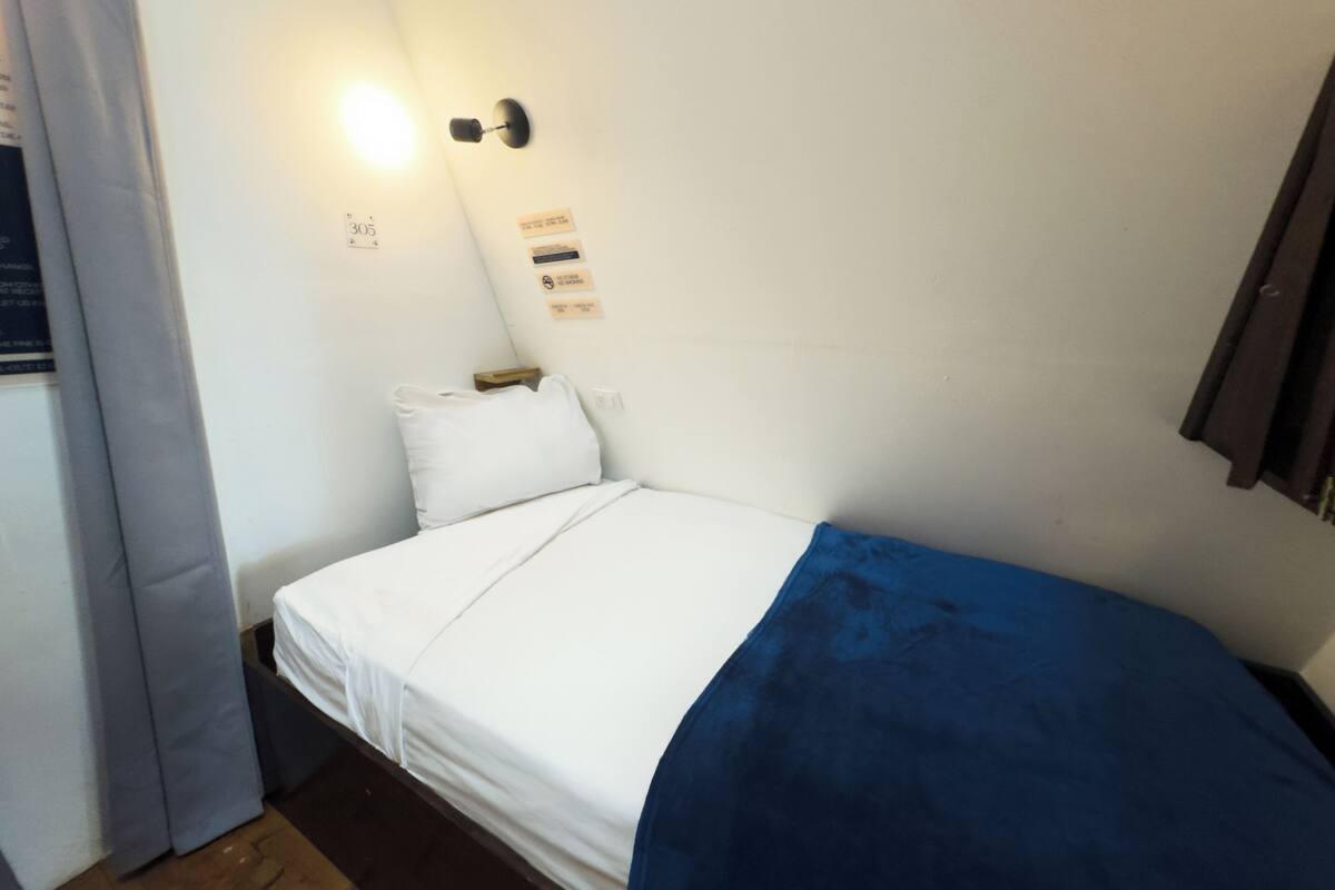Ein Einzelbett mit weißer Bettwäsche und einer blauen Decke, eine Wandleuchte und ein kleiner Nachttisch aus Holz mit einer Taschentuchbox.
