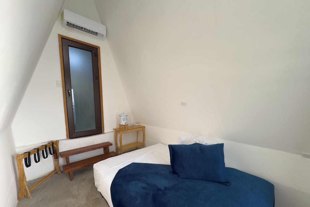 Ein kleiner, schlichter Raum mit einem Bett, einer hölzernen Bank, einem Tisch mit Lampe und einem Fenster mit Spiegel.