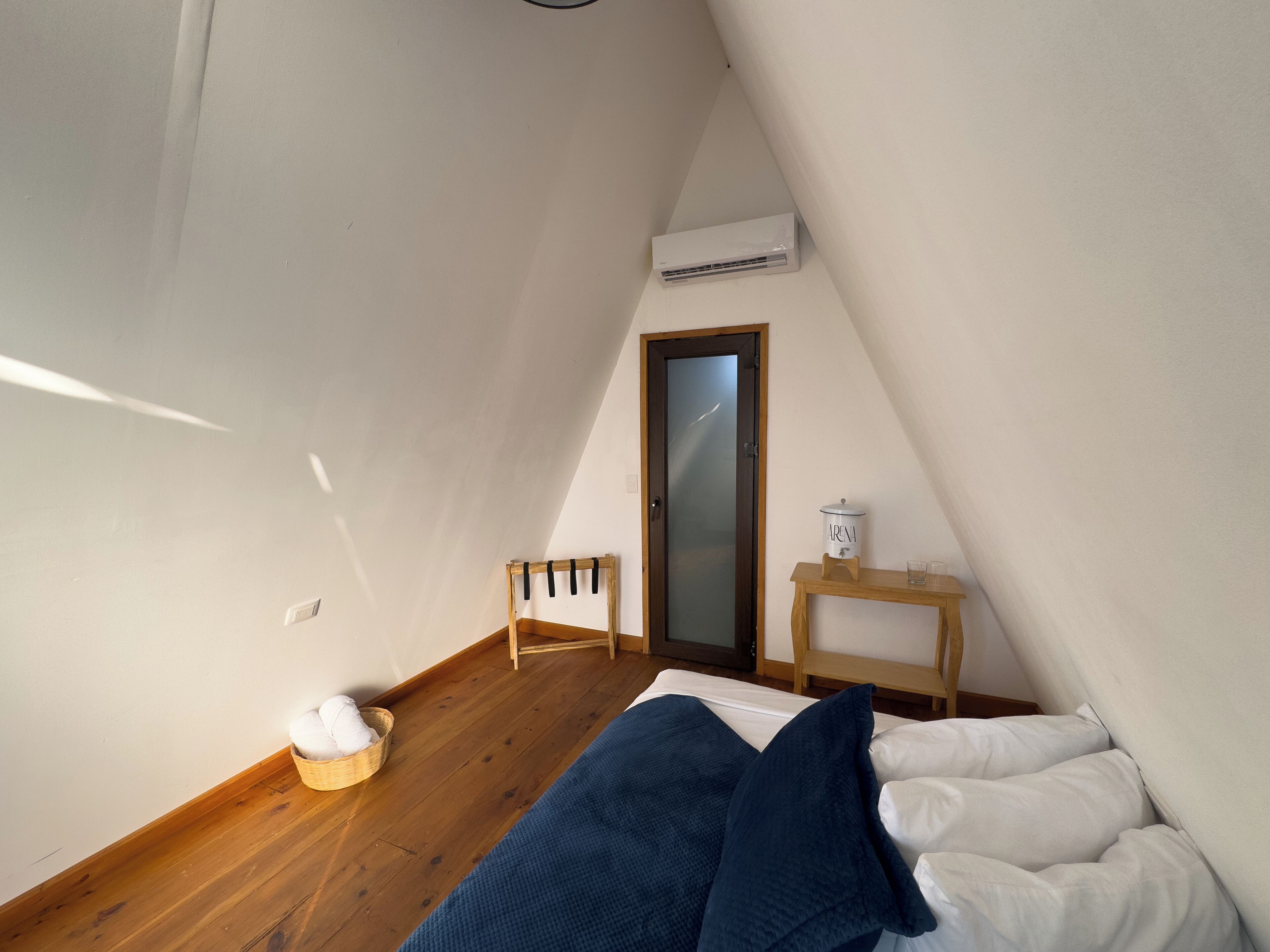 A small, cozy bedroom with a sloped ceiling, a wooden floor, a bed with white and blue bedding, a small wooden table with a lamp, and an air conditioning unit mounted on the wall.