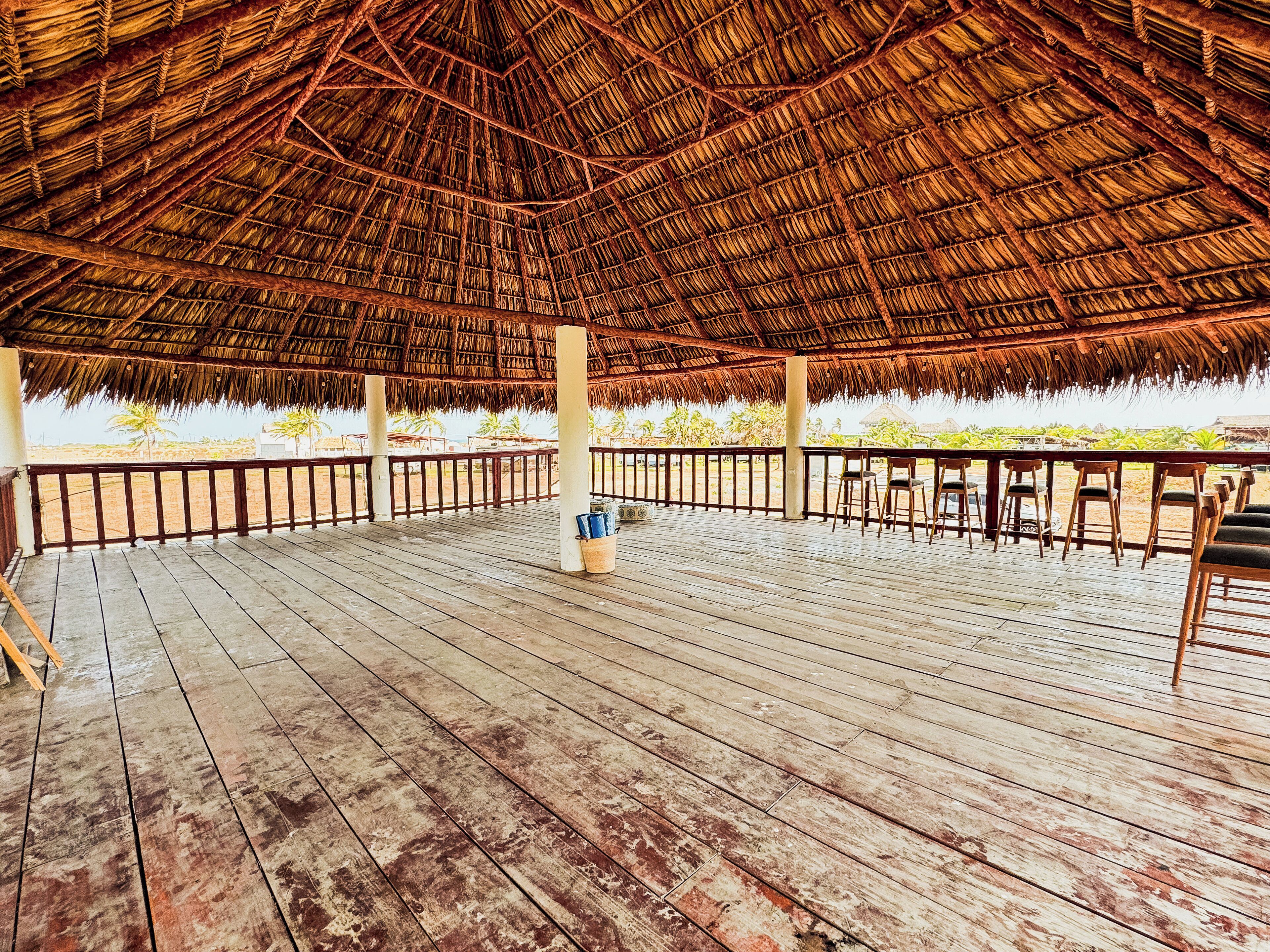 Una terraza de madera al aire libre con techo de paja, sillas de madera y vista a la playa.