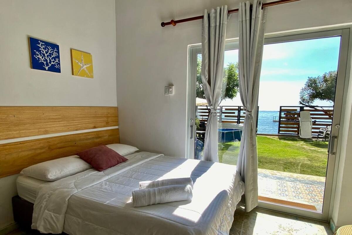 Une chambre à coucher avec un lit, une tête de lit en bois, une vue sur la mer et un balcon aménagé avec du mobilier d’extérieur.