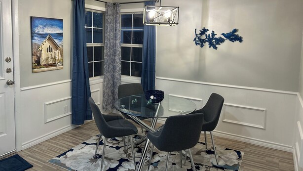 Un comedor con una mesa de vidrio, cuatro sillas, un adorno en la pared y una ventana con cortinas.