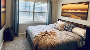 Un dormitorio con cama, ventilador de techo, ventana con cortinas y un cuadro en la pared.