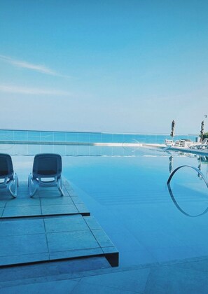 Un área de piscina con dos sillas reclinables mirando al agua, cielo despejado y vista al océano.