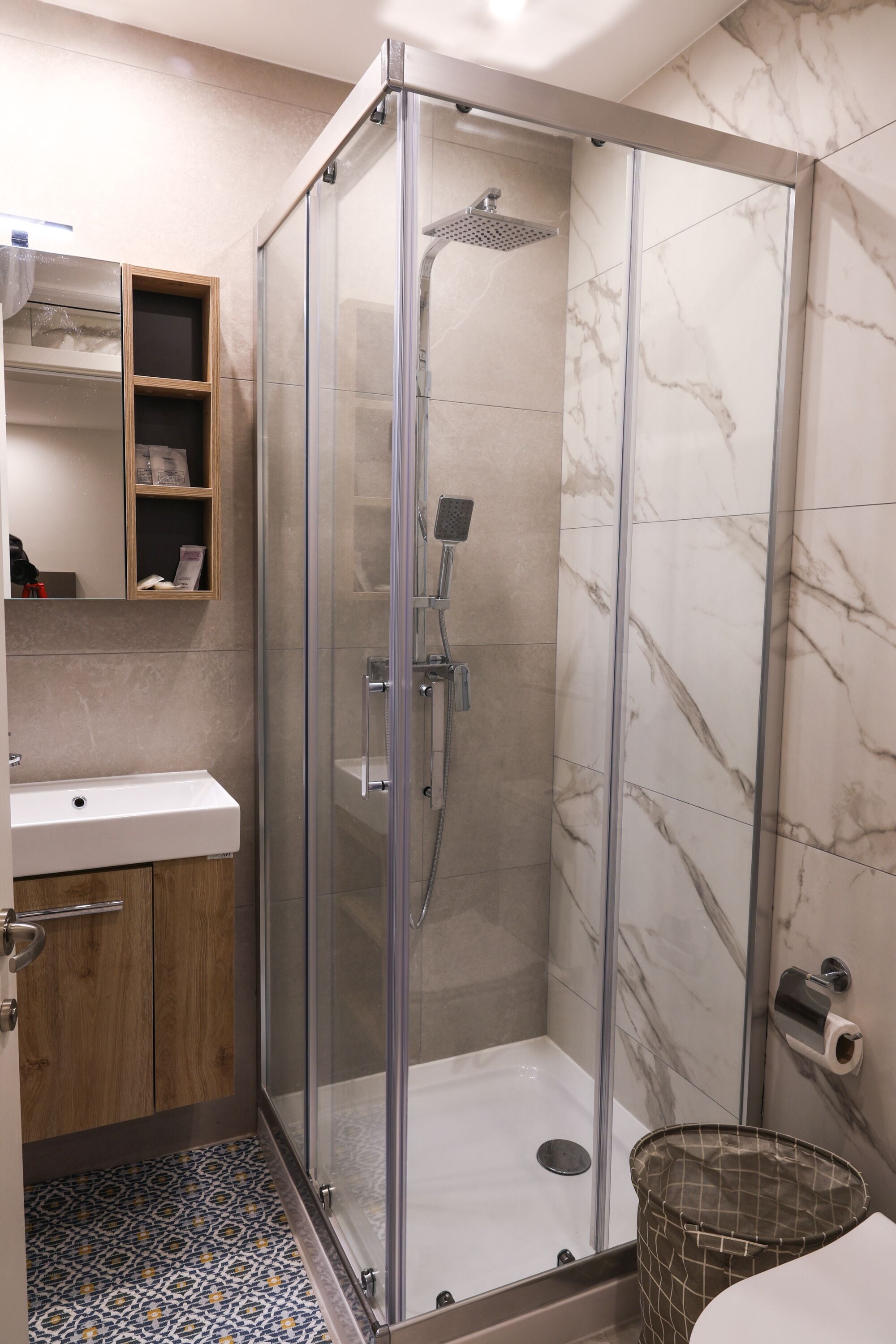 A modern bathroom with a glass-enclosed shower, a wooden vanity, and a patterned floor.