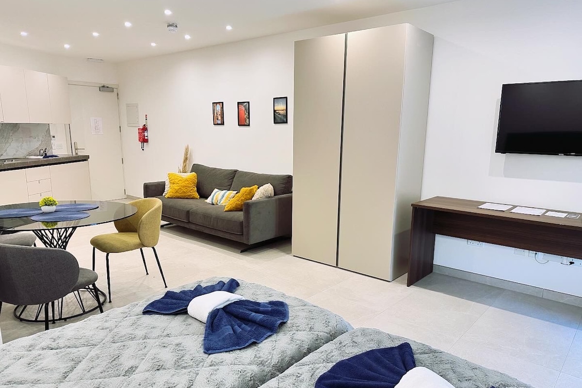 A modern hotel room with a sofa, two beds, a desk, a TV, and a kitchenette.
