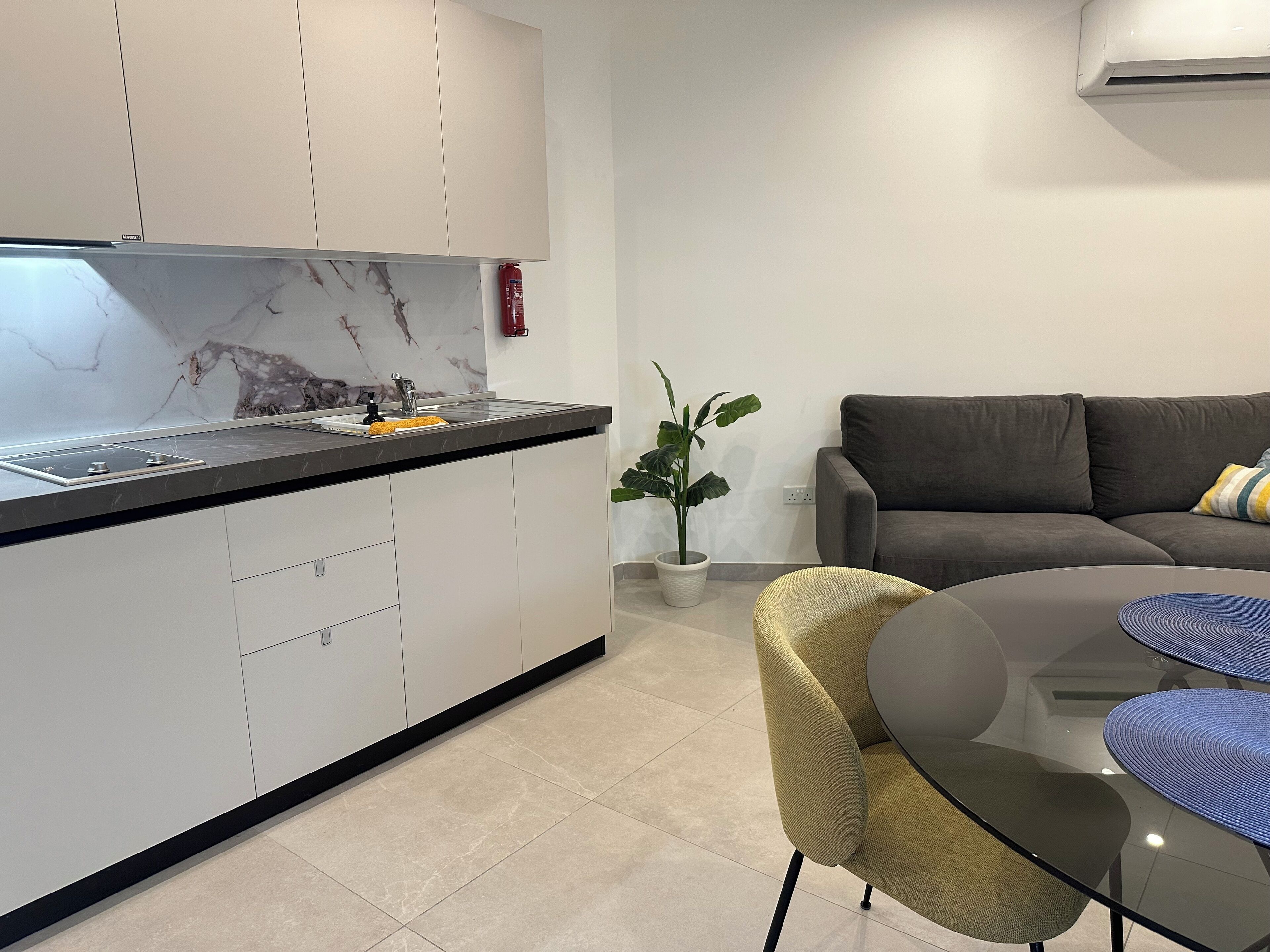 A modern kitchen with a marble backsplash, a built-in stove, and a sink. There is a dining area with a round glass table and chairs, a grey sofa, and a potted plant.