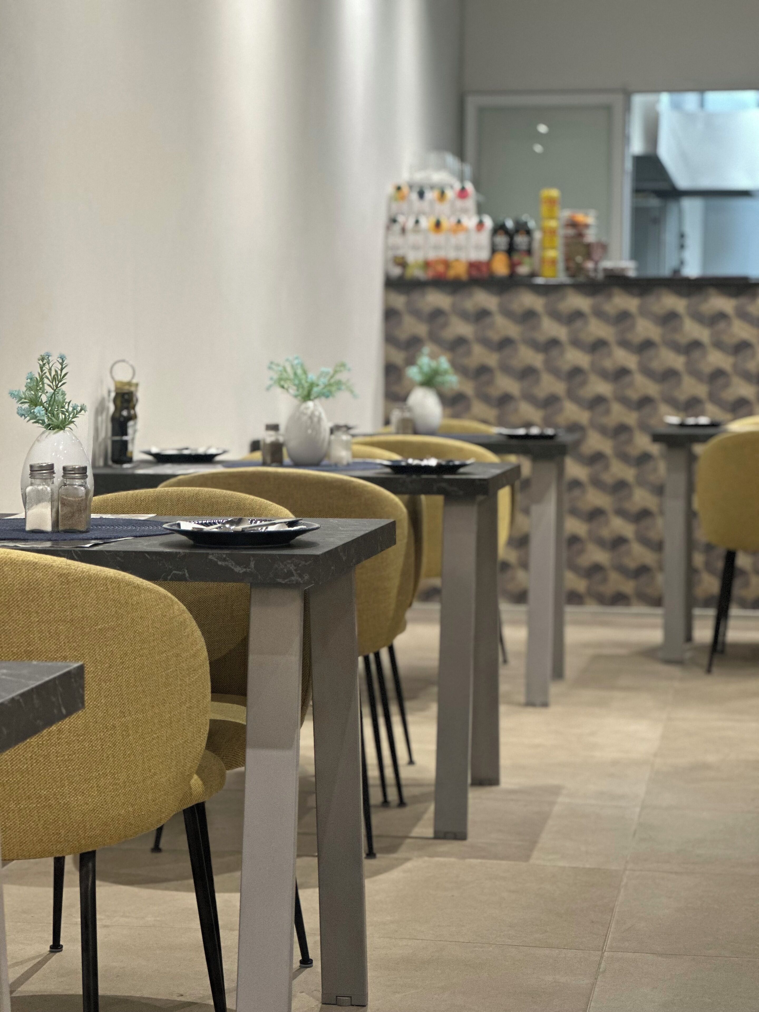 A modern dining area with yellow chairs and black tables, featuring a patterned wall and a kitchen in the background.