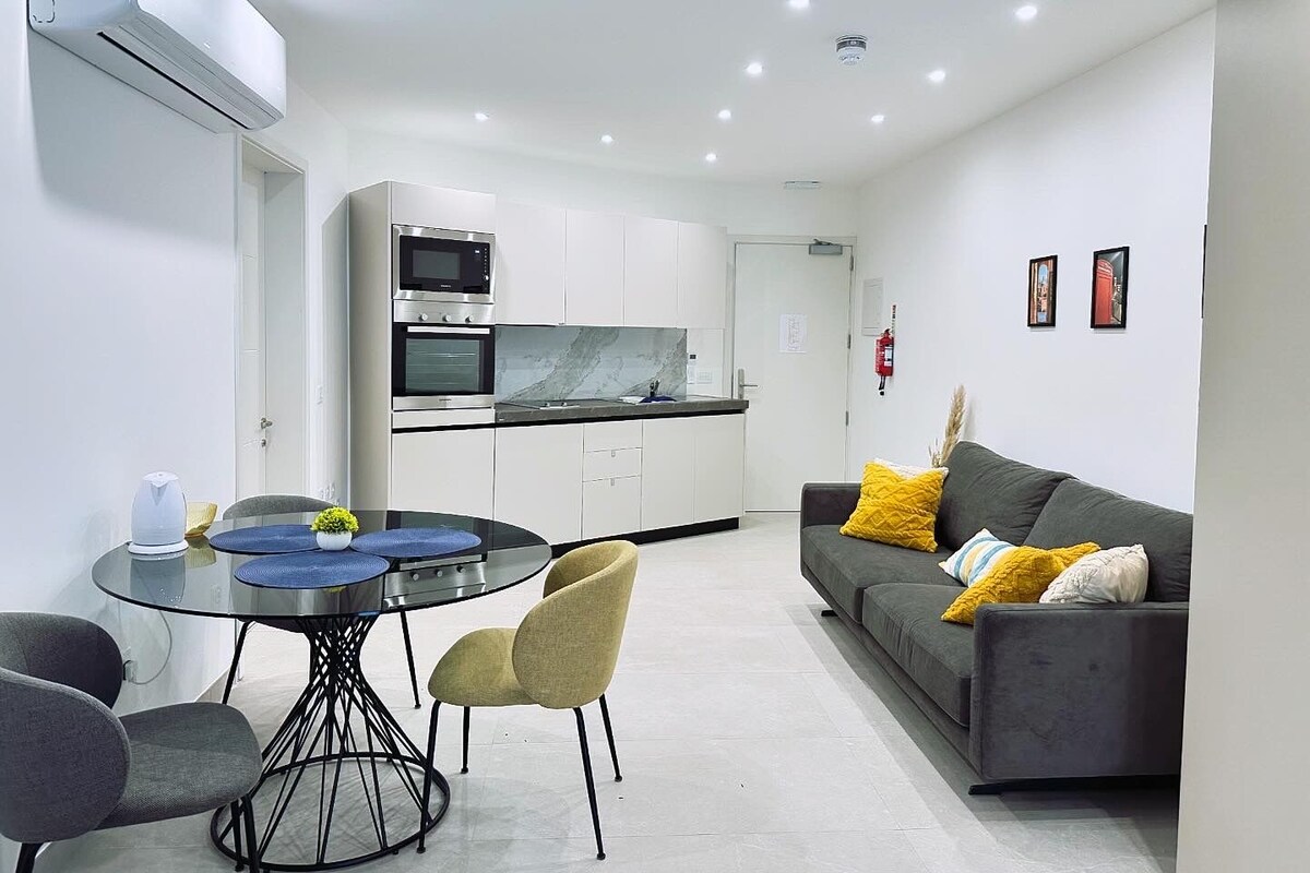 Una cocina moderna con electrodomésticos integrados, una zona de comedor con una mesa redonda y sillas, y un sofá gris con cojines amarillos.