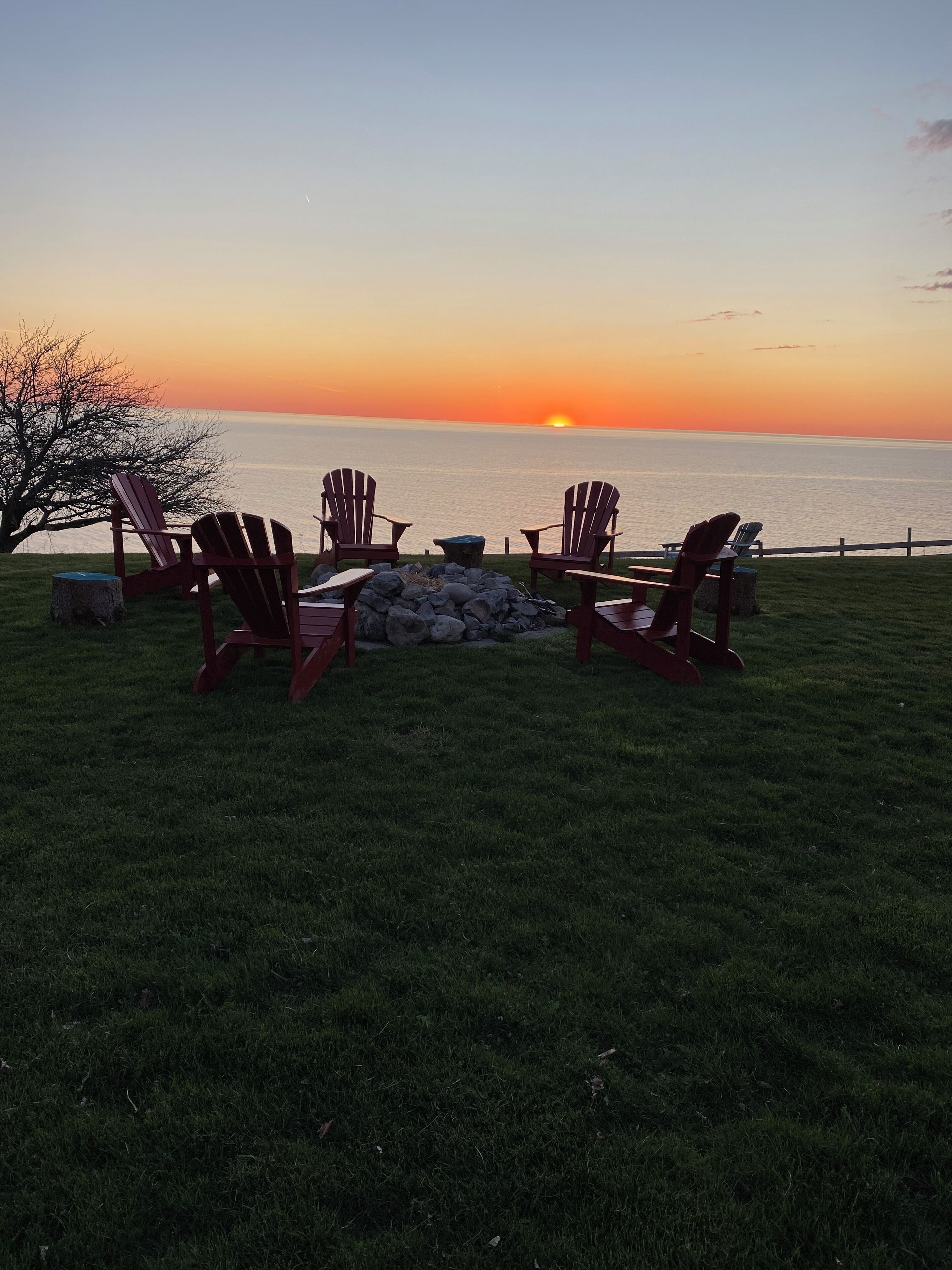 A sunset view with chairs and a fire pit on a grassy area by the water.
