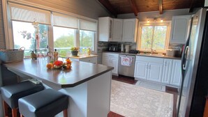 Una cocina moderna con armarios blancos, una isla central con taburetes y un refrigerador, y una ventana con vista a árboles.