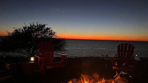 Vista al atardecer con una fogata y dos sillas Adirondack junto al agua.