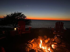 Vista do pôr do sol com lareira e duas cadeiras Adirondack à beira da água.