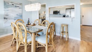 Un coin repas avec une table en verre et des chaises en osier, un lustre, et une cuisine visible à travers une porte ouverte.