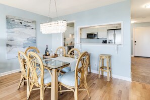 Zona pranzo con tavolo in vetro e sedie in vimini, un lampadario e una cucina visibile attraverso un'apertura.