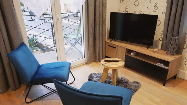 Sala de estar com duas cadeiras azuis, uma TV sobre um móvel de madeira e uma mesa redonda pequena com um controle remoto.