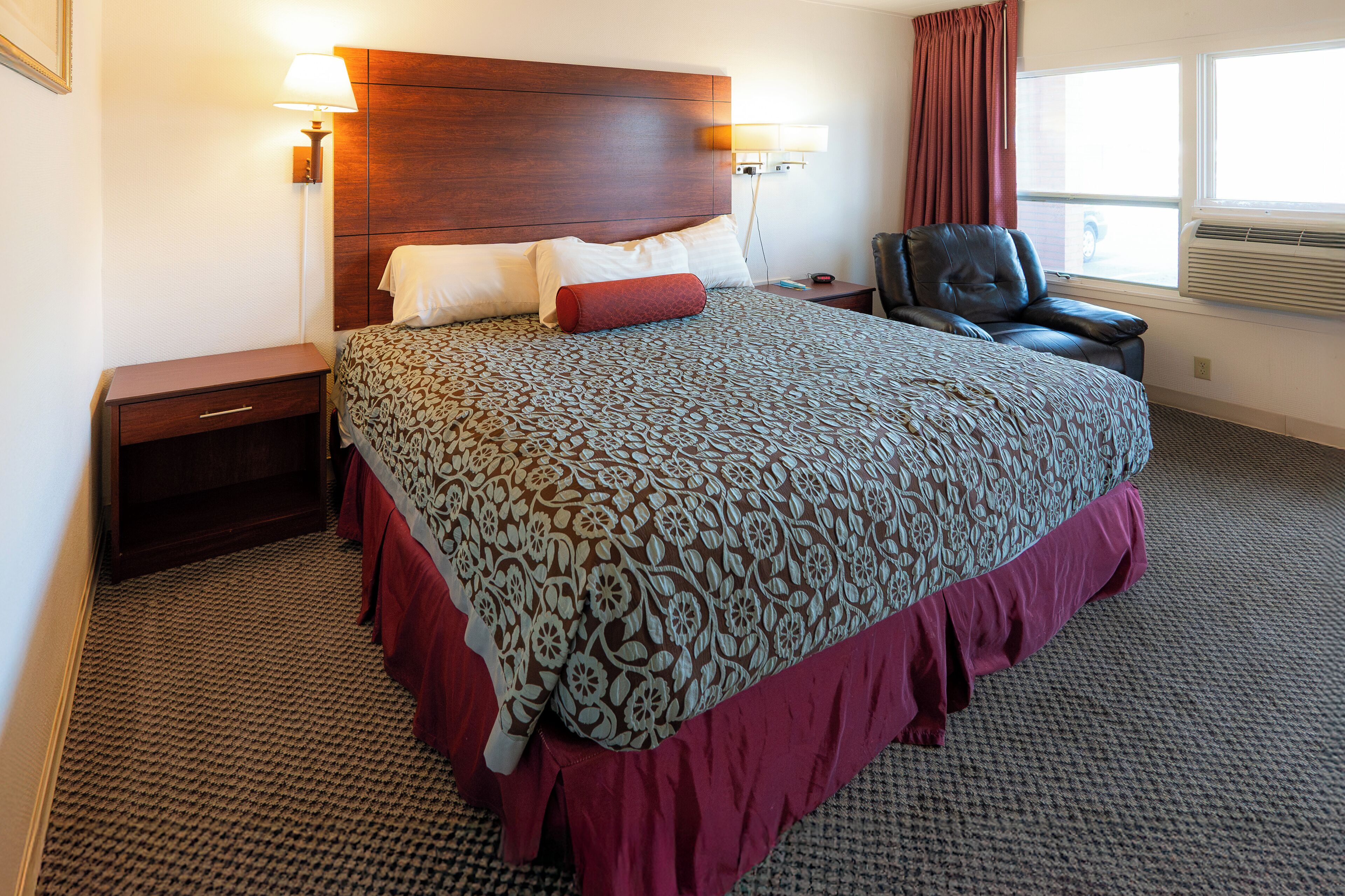 A hotel room with a large bed, a wooden headboard, a nightstand, a chair, and a window with curtains.