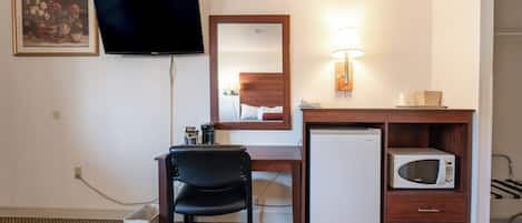 A hotel room with a desk, a television, a refrigerator, a microwave, a chair, and a bedside lamp.