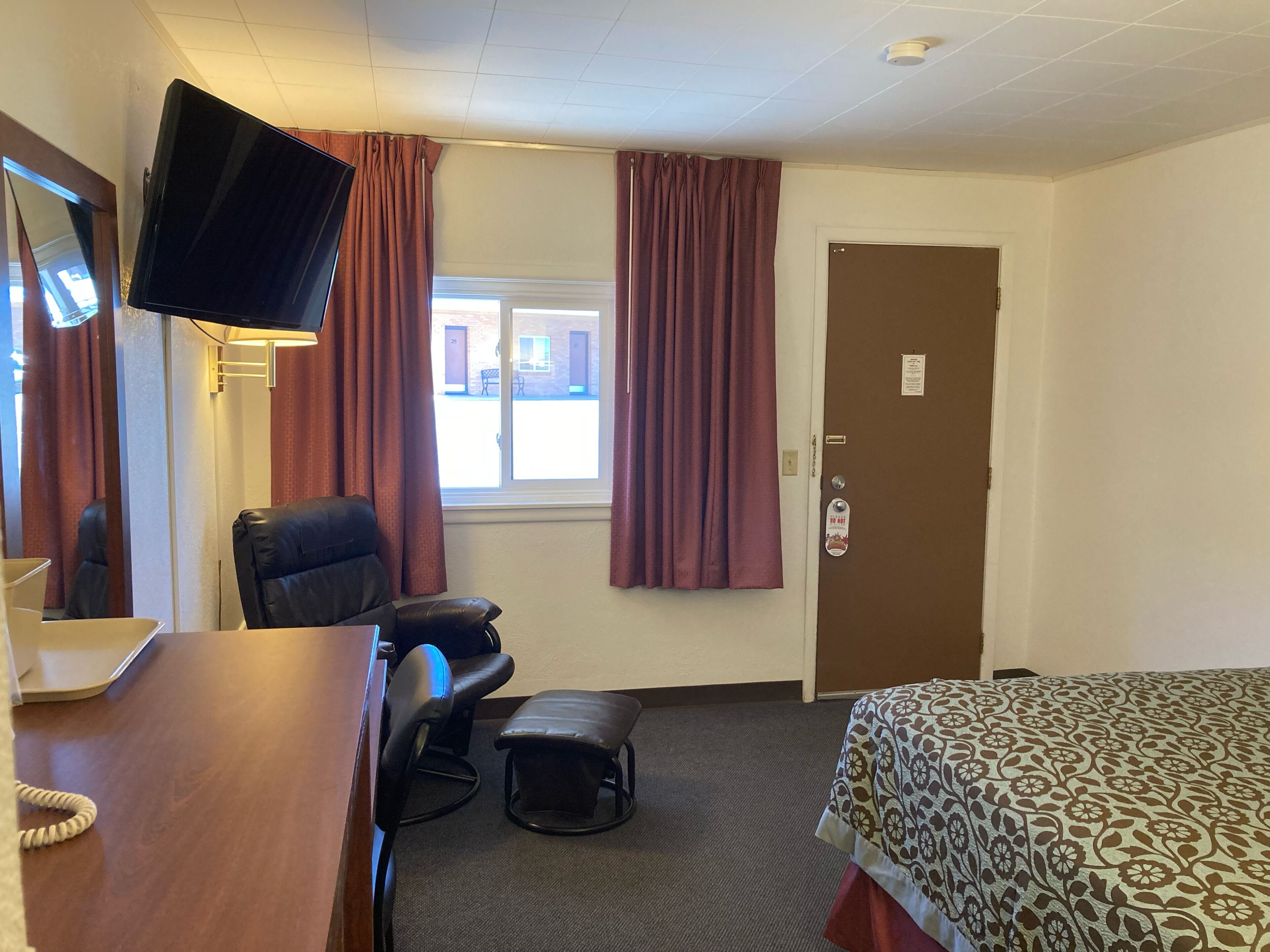 A hotel room with a bed, a desk with a telephone, a chair, an ottoman, a television mounted on the wall, and a door with a lock.