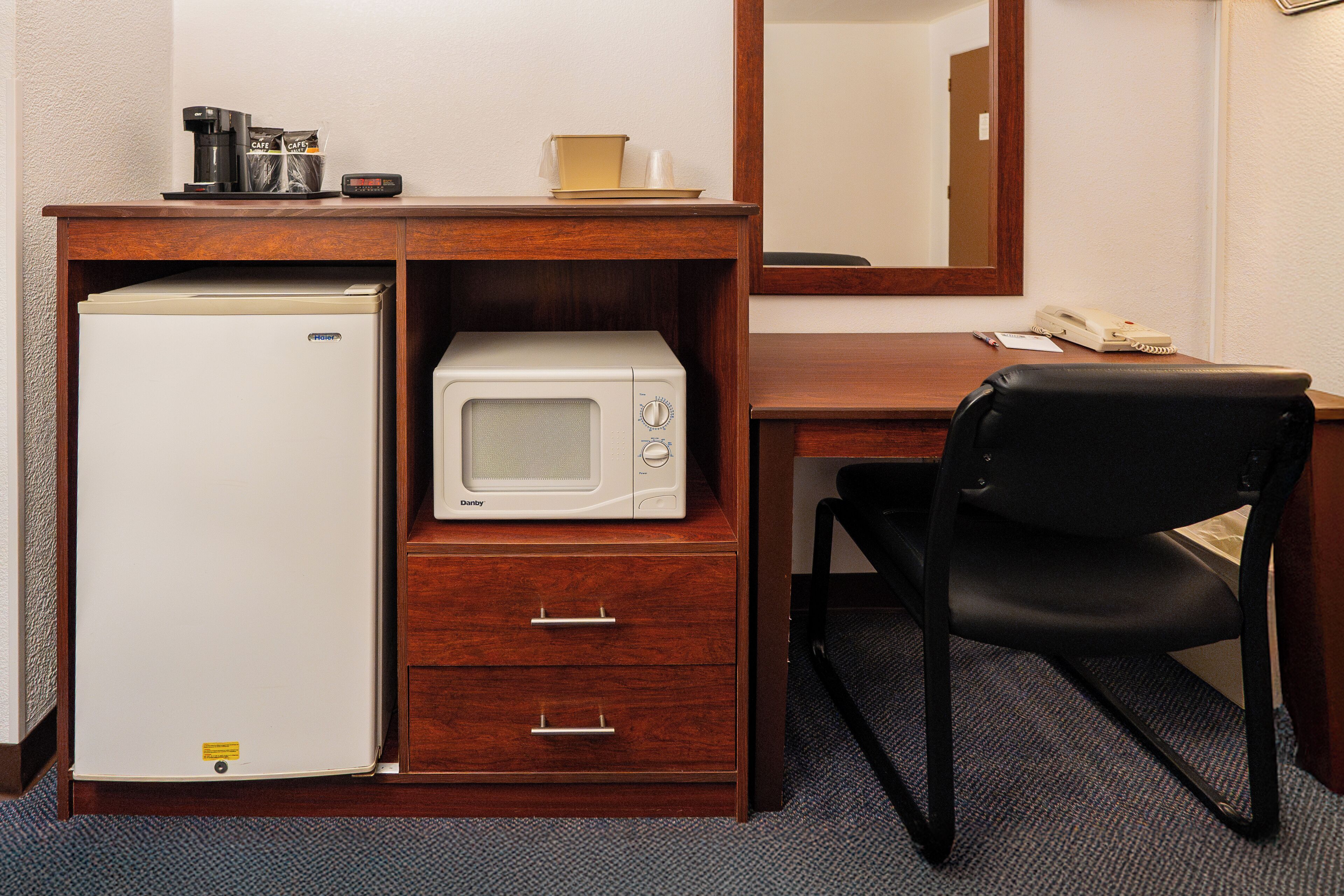 Ett kompakt hotellrum med ett skrivbord i trä, en svart kontorsstol, ett vitt kylskåp, en mikrovågsugn och en kaffebryggare.