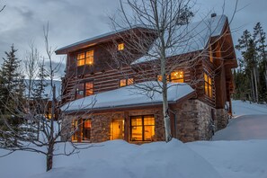 Una cabaña de troncos de varios pisos con base de piedra, ventanas grandes y techo cubierto de nieve.