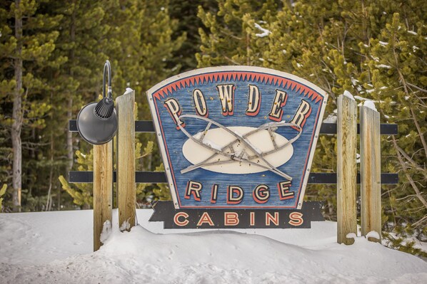 Une enseigne indiquant l’emplacement des chalets de Powder Ridge, avec une clôture en bois rustique et un sol recouvert de neige.