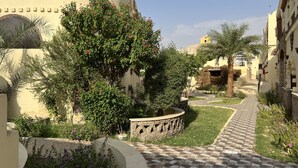 Un patio con un camino empedrado, rodeado de vegetación y un edificio con muro de piedra.
