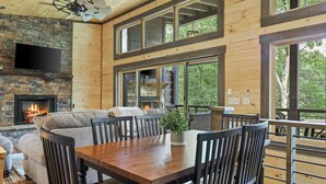 Een knus chaletinterieur met een stenen openhaard, een groot raam met uitzicht op een bos, en een eethoek met een houten tafel en stoelen.