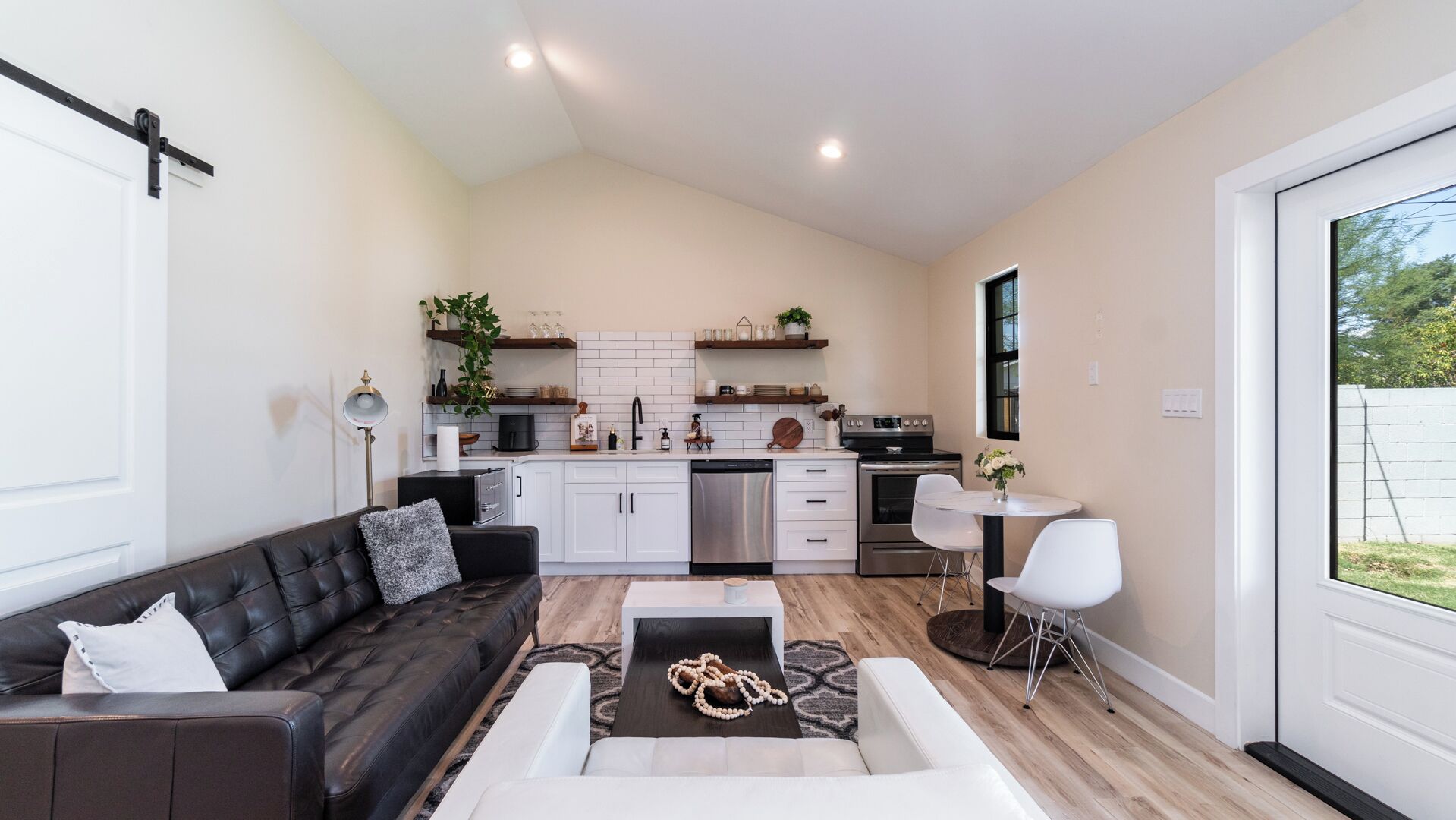 A modern kitchen with white cabinets, a black countertop, and stainless steel appliances. There is a dining area with a white table and chairs, and a living room with a black leather sofa and a coffee table.