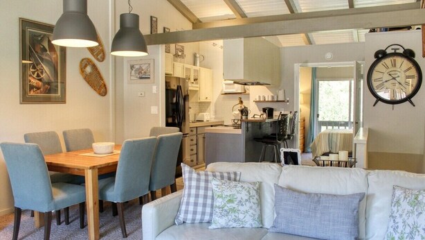 Un salon confortable avec un canapé, une table à manger et des chaises. Il y a un coin cuisine avec des placards et un réfrigérateur. Une horloge est fixée au mur.