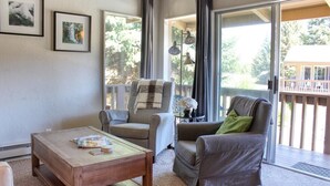 Un salon chaleureux avec une table basse en bois, deux fauteuils et un canapé. Des tableaux encadrés ornent le mur, et une grande fenêtre est agrémentée de rideaux.