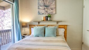 Une chambre à coucher avec un lit, une fenêtre avec des rideaux, une étagère fixée au mur avec des livres et une plante, et un grand tableau floral au-dessus du lit.