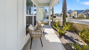 Een overdekt terras met rieten stoelen en een glazen tafel, met uitzicht op een straat met palmbomen en huizen.
