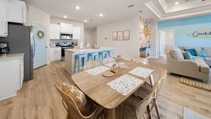 Een moderne keuken met witte kastjes, een inbouwoven en een koelkast. Er is een eethoek met een houten tafel en rieten stoelen. Een blauwe deur leidt naar een andere kamer.