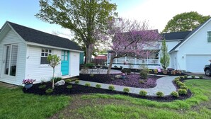 A residential property with a small house, a garden with a paved walkway, and a large tree with purple blossoms. - Quaint historic home in the heart of Bemus Point Village- a stroll to the water! (Bemus Point)