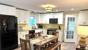 A modern kitchen with white cabinets, a black refrigerator, a microwave, and a stove. There is a dining table with wooden chairs and a bench. - Quaint historic home in the heart of Bemus Point Village- a stroll to the water! (Bemus Point)