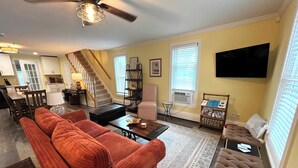 A living room with a sofa, coffee table, and ottoman, a staircase leading to a kitchen, and a flat-screen TV mounted on the wall. - Quaint historic home in the heart of Bemus Point Village- a stroll to the water! (Bemus Point)