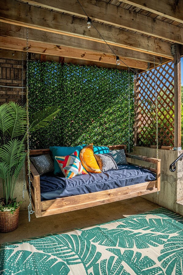 Un lit-balançoire en bois avec un matelas bleu, entouré d’une végétation luxuriante et agrémenté d’un tapis à motifs.